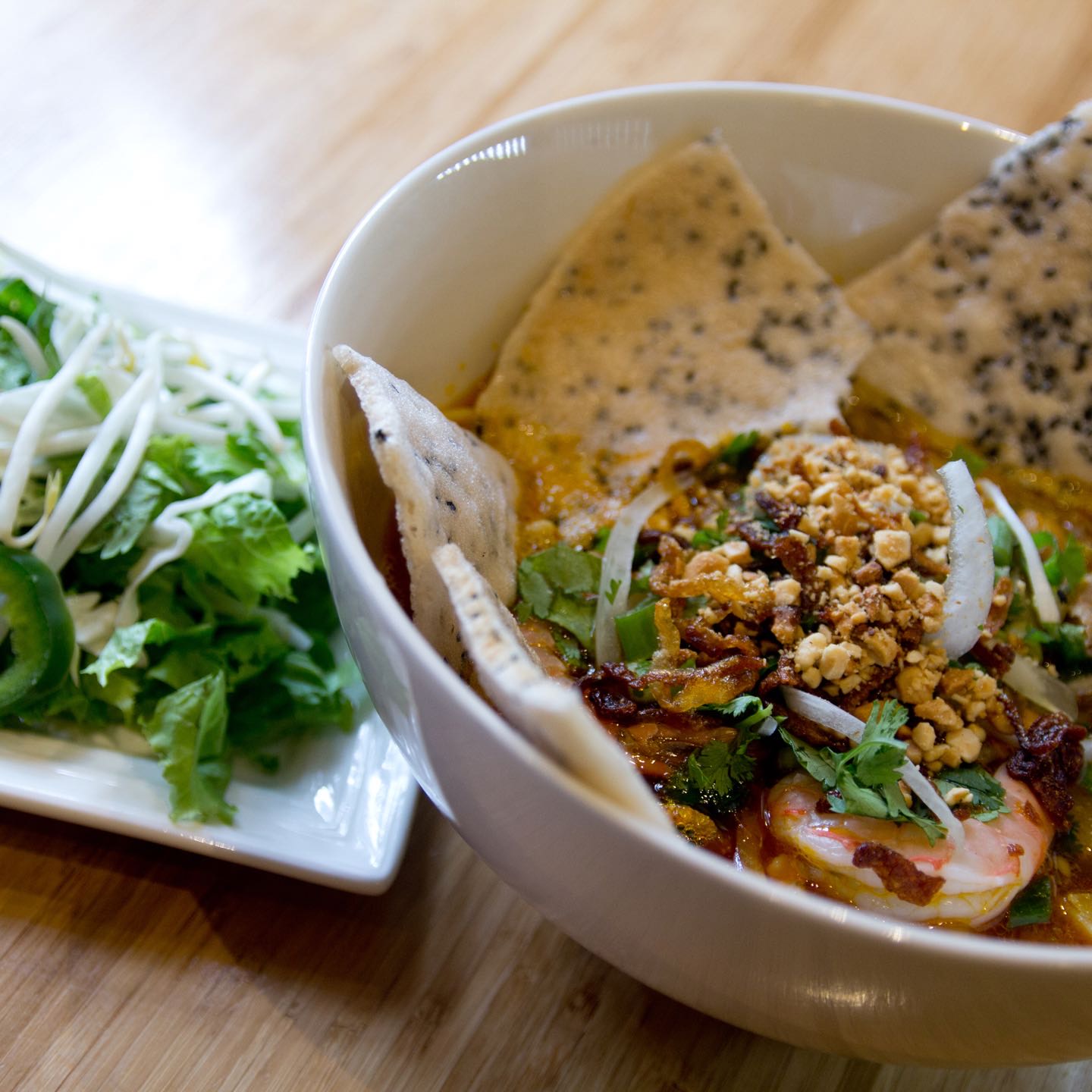 Seasoned turmeric broth ladled over wide rice noodles, minced pork, shrimp and diced jicama topped with roasted peanuts, served with sesame rice crackers.